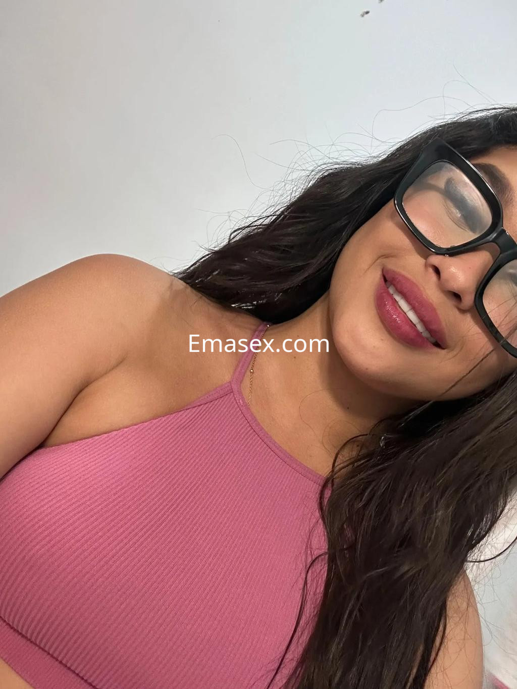 Escort en Granada con cabello largo y oscuro, usando un top rosa y gafas, sonríe ligeramente hacia la cámara.