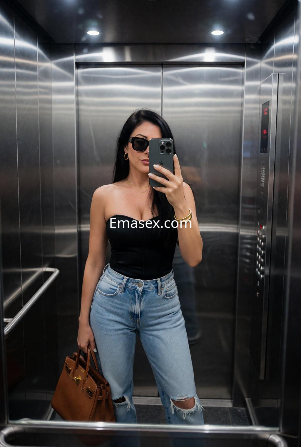 Escort en Málaga con un body negro y jeans claros, posando en un ascensor con gafas de sol y un bolso marrón.