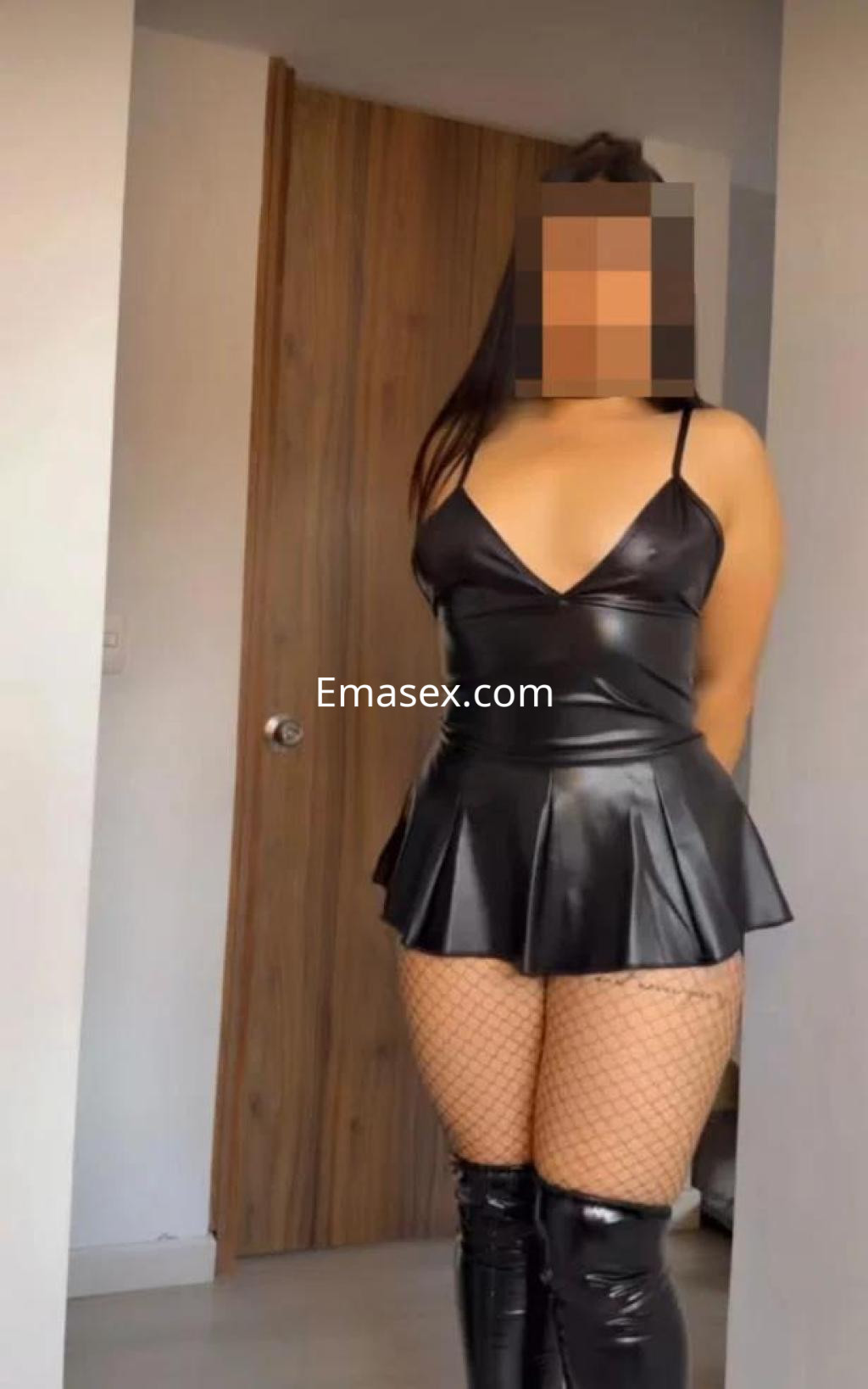 Escort en Toledo, vistiendo un ajustado vestido de cuero negro con tirantes y medias de red, en un entorno interior.