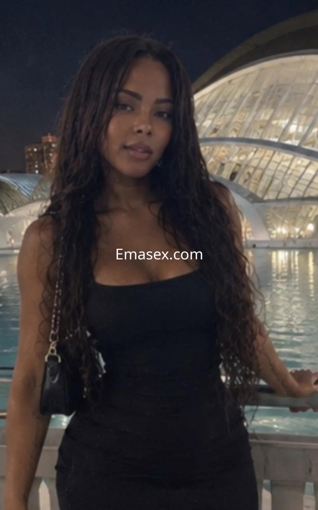 Puta de cabello largo y rizado, viste un vestido negro ajustado, posando en Valencia junto a un moderno edificio.