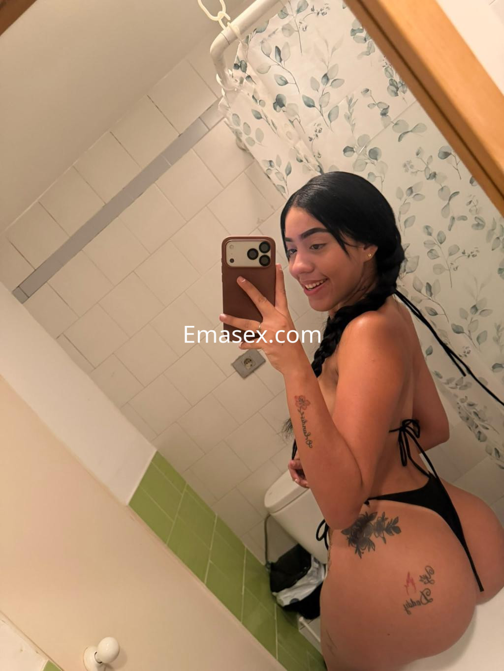 Escort posando frente a un espejo en un baño, luciendo un bikini negro y mostrando tatuajes en la piel en Madrid.
