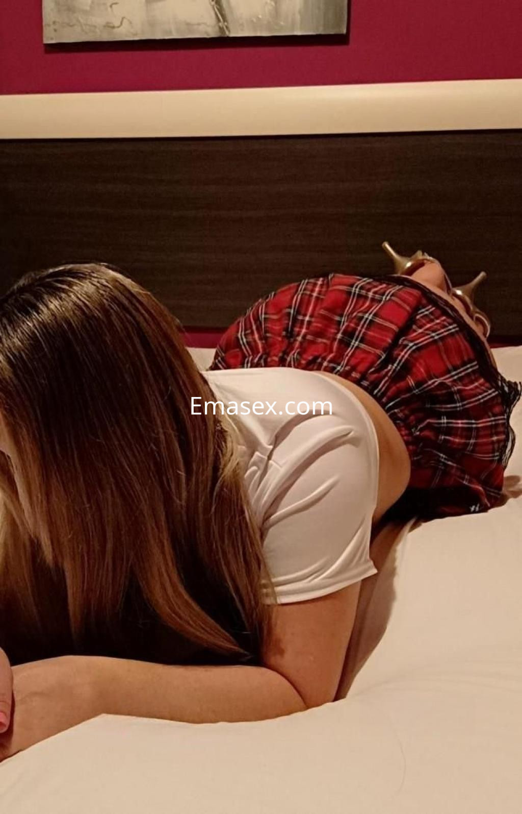 Escort en Vigo, recostada sobre la cama, luciendo una blusa blanca y una falda corta de cuadros rojos.