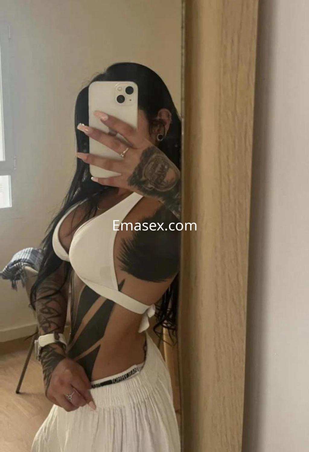 Escort en Palma de Mallorca, posando frente al espejo con un top blanco y pantalones cortos, mostrando tatuajes.
