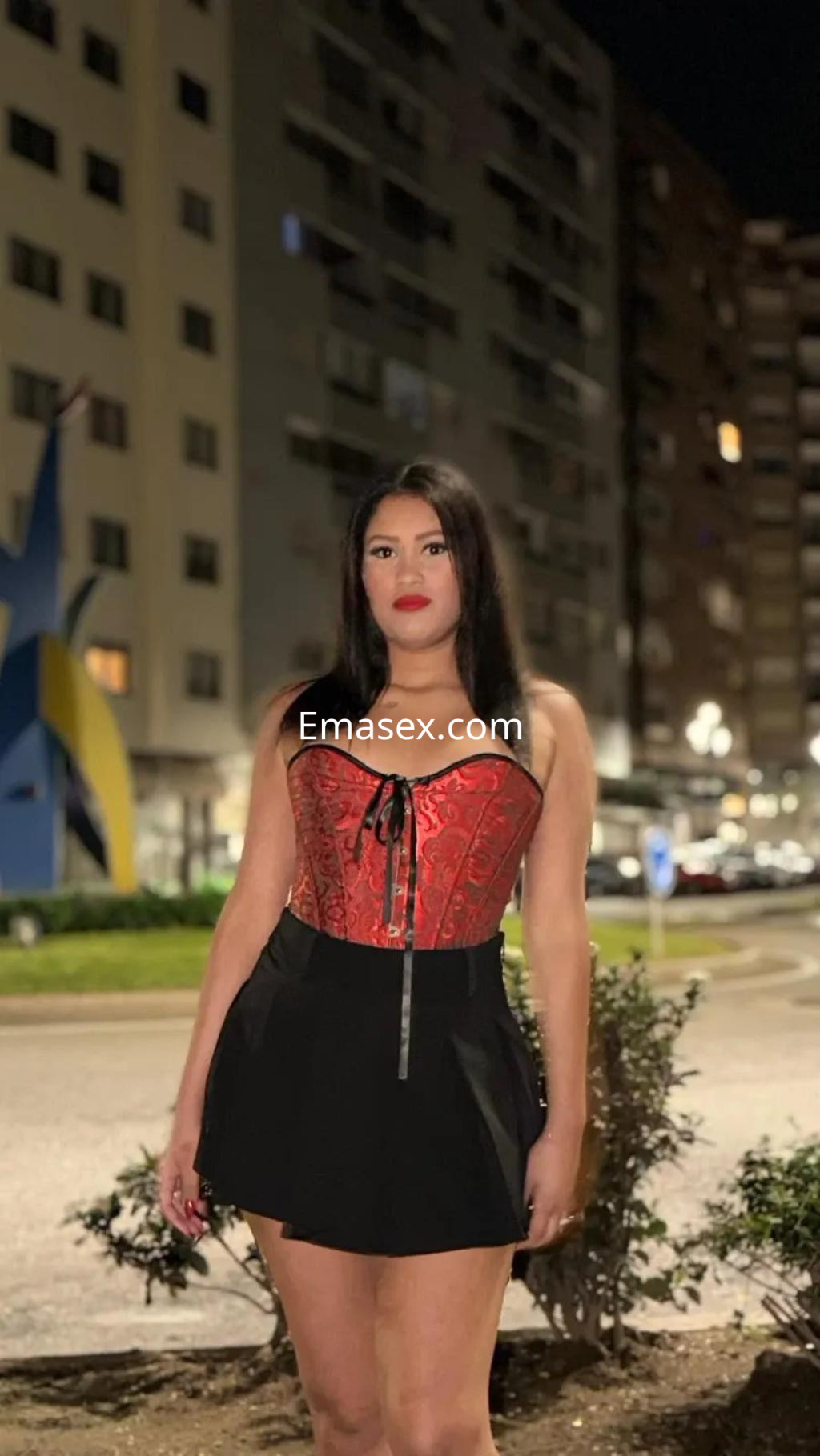 Puta en Vigo, luciendo un corsé rojo con detalles oscuros y una falda corta negra, en un entorno urbano nocturno.