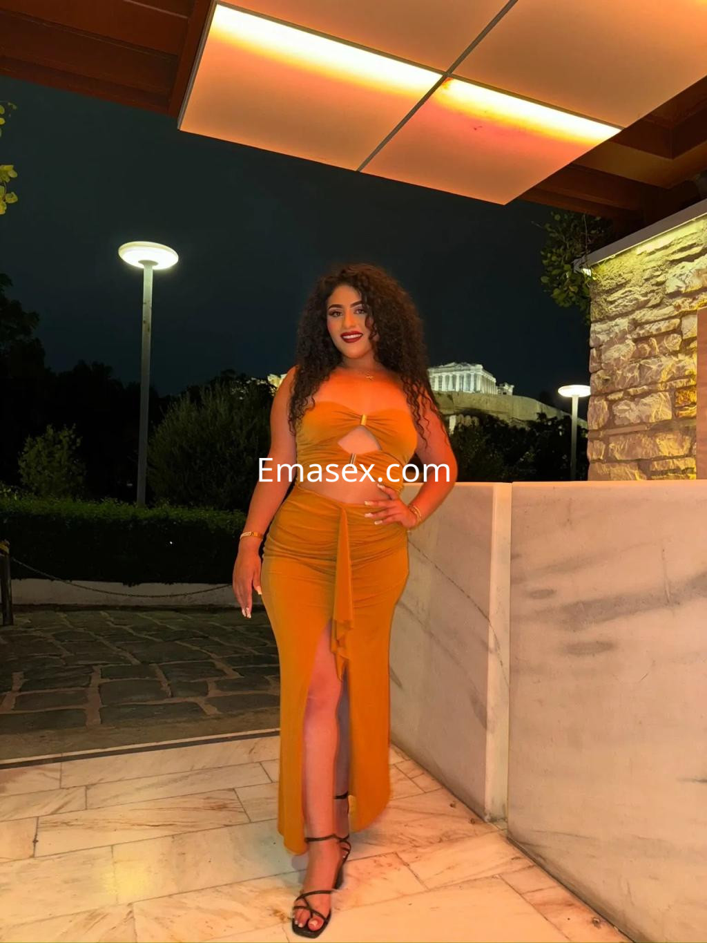 Puta en Palma de Mallorca, luciendo un vestido largo naranja con abertura y sandalias, posando en un ambiente elegante.