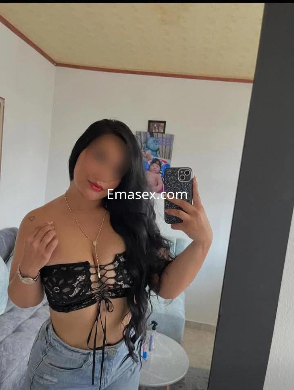 Escort en Córdoba, con cabello largo y oscuro, viste un top negro de encaje y jeans, posando frente a un espejo.