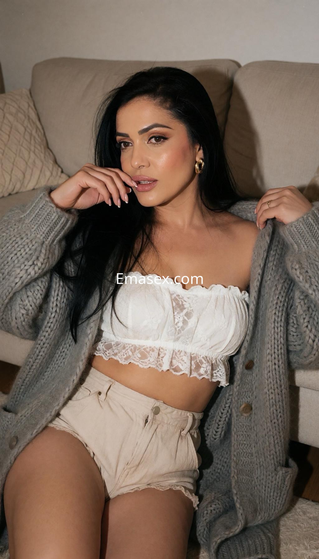 Escort en Pamplona, sentada en un sofá con un top blanco de encaje y un cardigan gris, mostrando una expresión seductora.