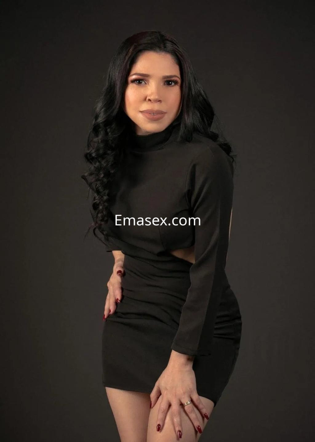 Escort en Oviedo con cabello largo y negro, vistiendo un vestido negro ajustado y posando de manera seductora.