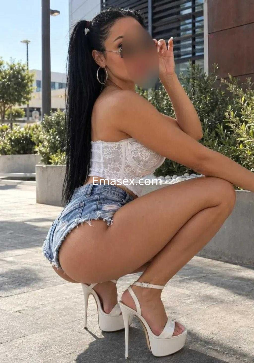 Escort en Toledo, en una pose agachada con blusa de encaje y shorts de mezclilla, luciendo tacones altos.