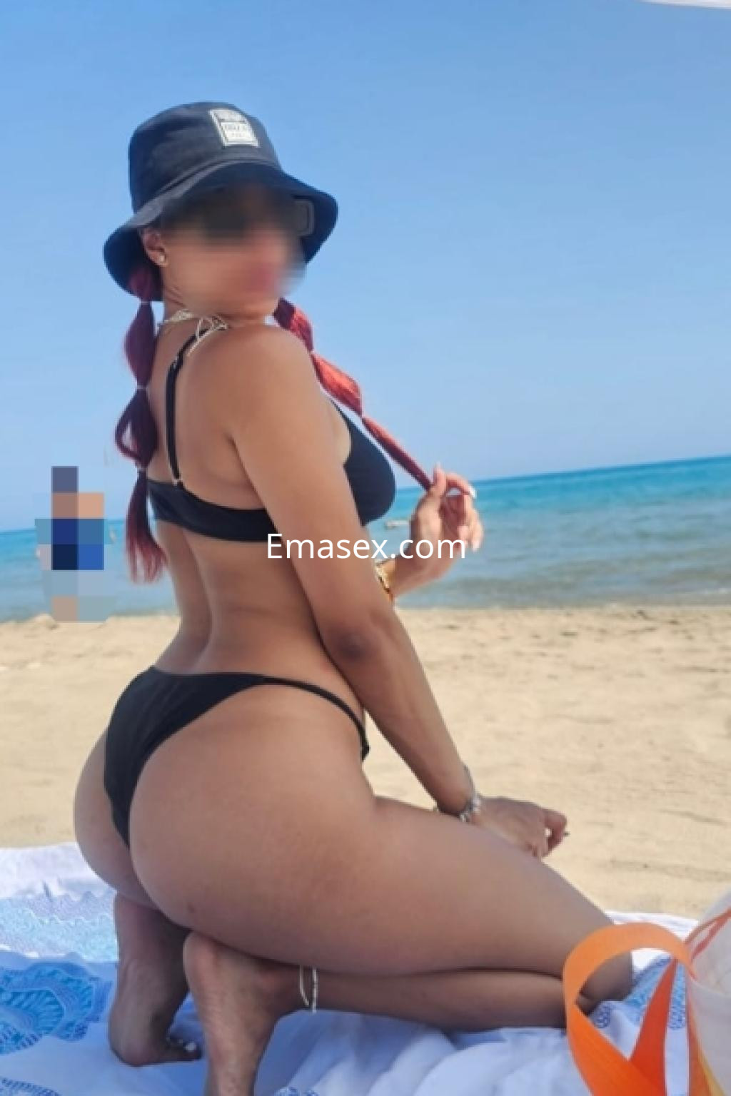 Puta de Almería posando en la playa con un bikini negro y un sombrero, mostrando su figura en un entorno soleado.