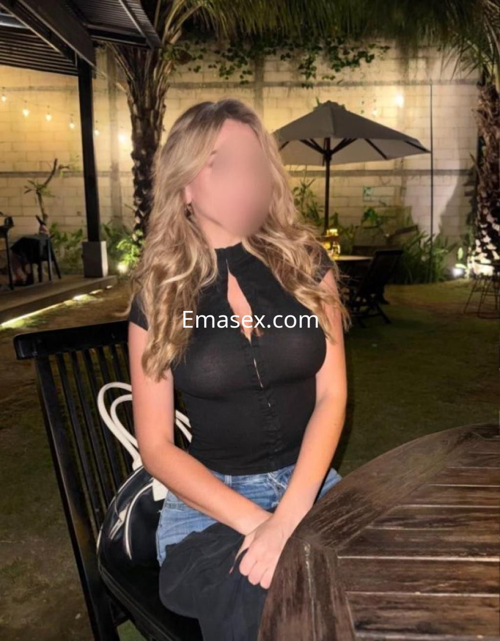 Escort en Almería sentada en una mesa al aire libre, vistiendo una blusa negra y jeans, con cabello rubio.
