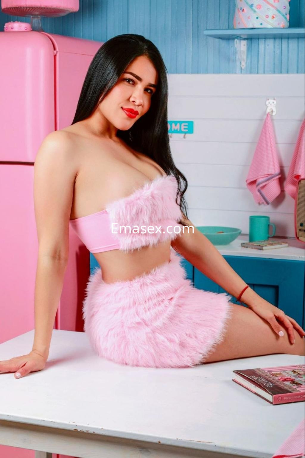 Escort en Bilbao posando con un conjunto rosa de pelaje, sentada sobre una mesa en una cocina colorida.