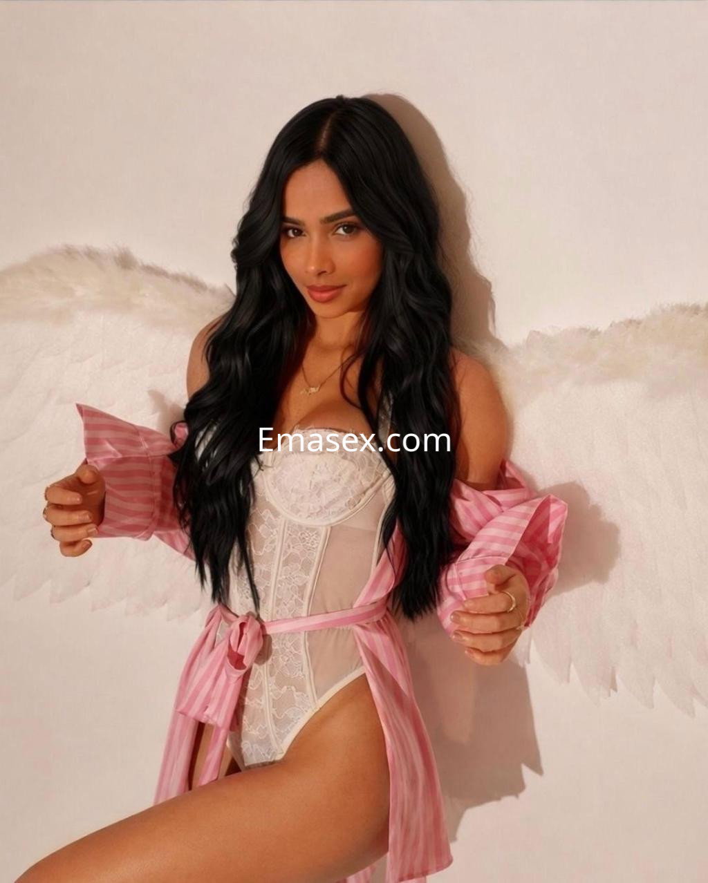 Escort en Valladolid posa con alas blancas, vistiendo un body blanco y una bata rosa a rayas, con cabello largo y oscuro.