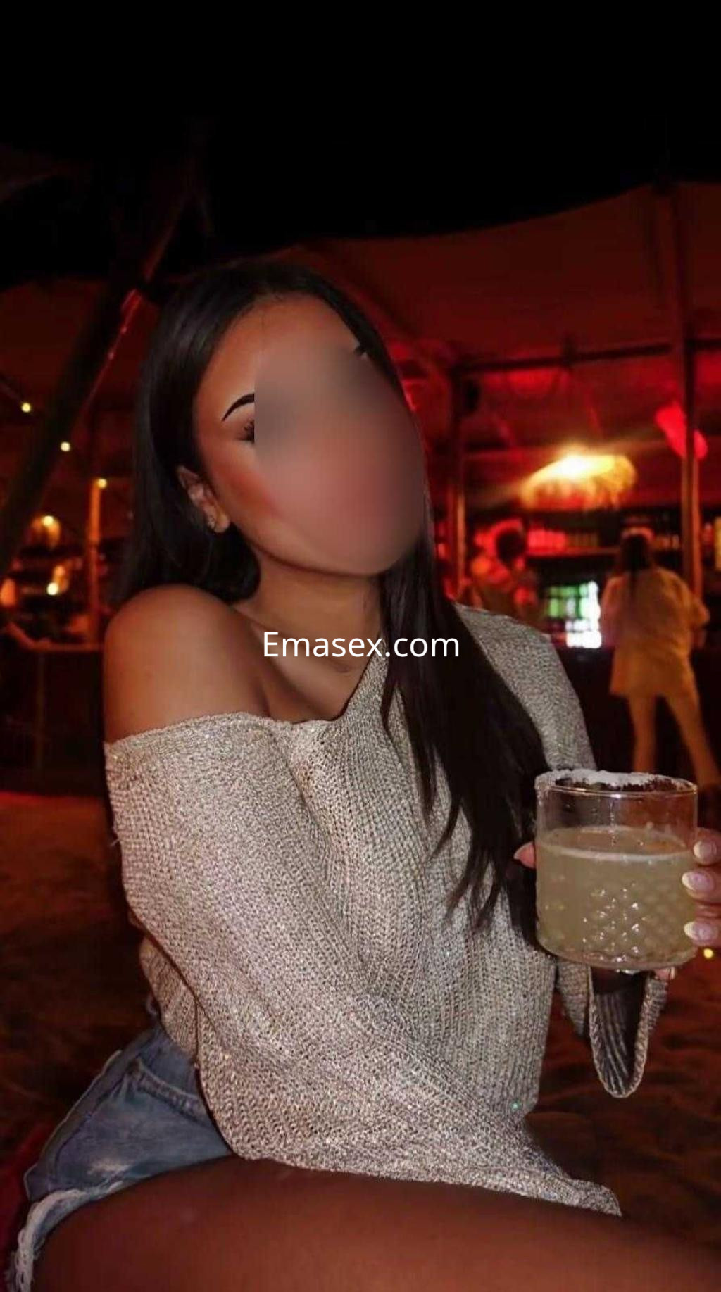Escort en Madrid, con top de manga larga y shorts, sosteniendo una bebida en un ambiente de bar iluminado.