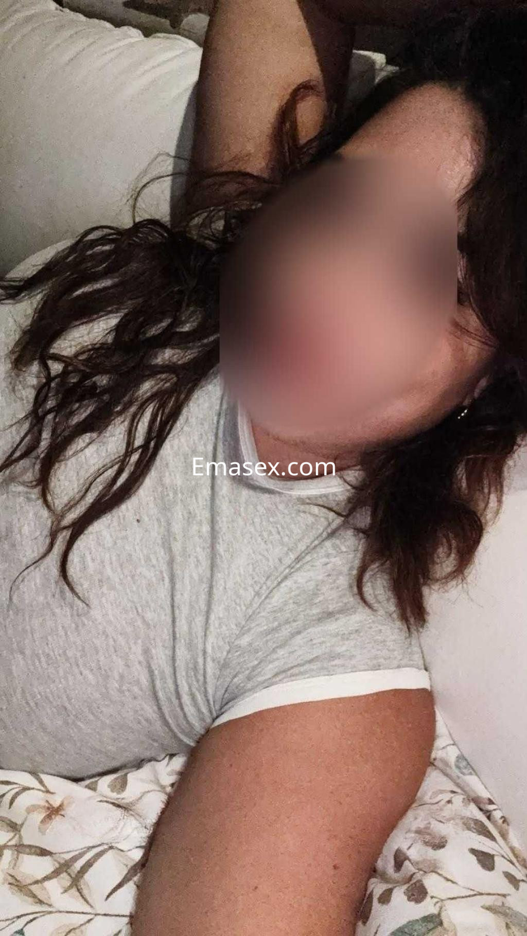 La puta en Córdoba posa recostada sobre una cama, vistiendo una camiseta gris y mostrando su cabello rizado.
