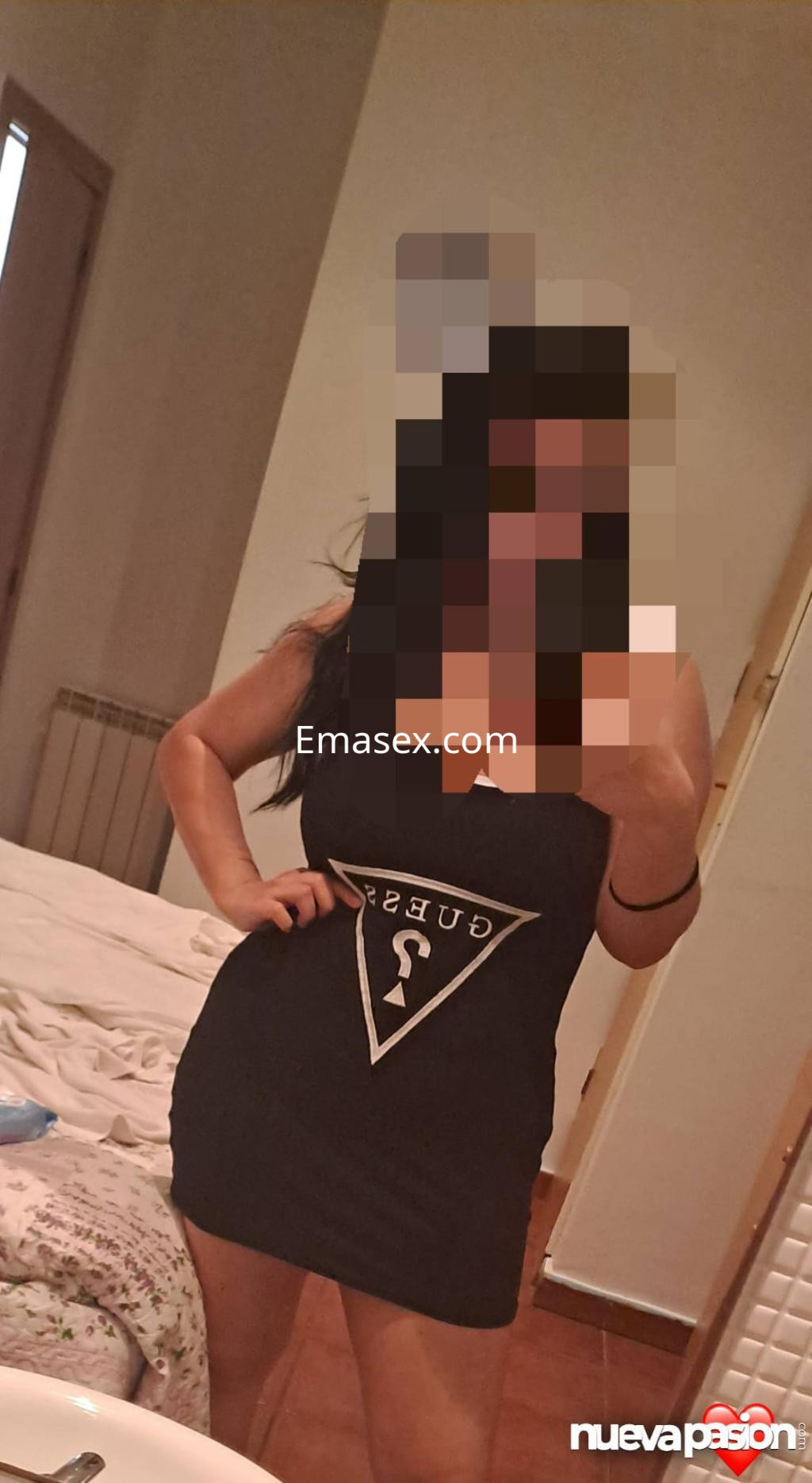 Escort en Lleida luce un vestido negro ajustado con un logo en el pecho, posando en un ambiente doméstico.