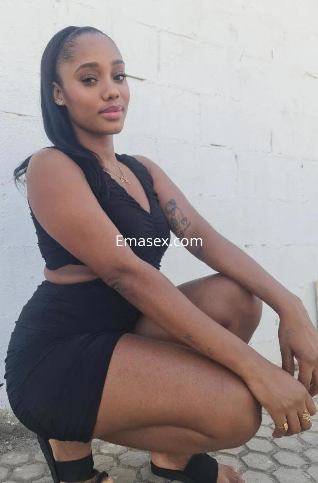 Escort en Sevilla, con vestido negro ajustado, posando de rodillas y luciendo un peinado lacio y elegante.