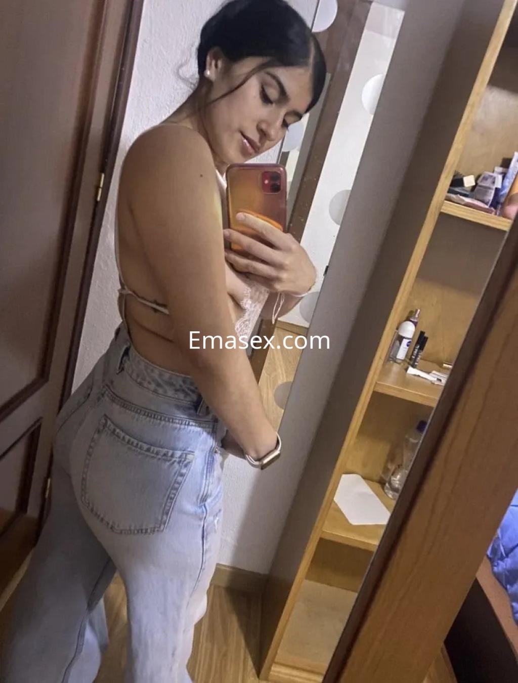 Puta en Córdoba posando de perfil con blusa sin mangas y jeans ajustados frente a un espejo.