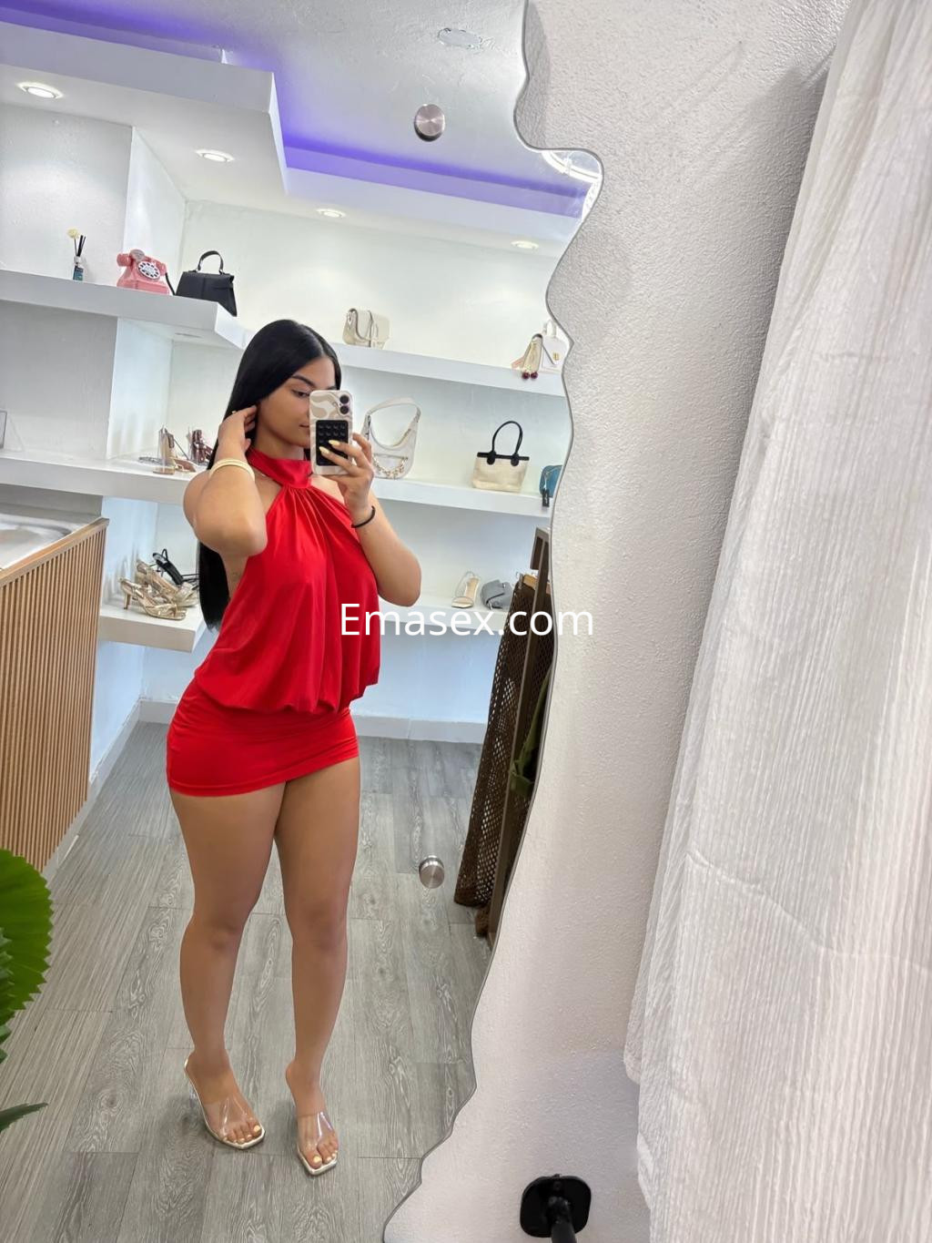 Escort en Zaragoza, luciendo un vestido rojo ajustado y sandalias transparentes, posando frente a un espejo.