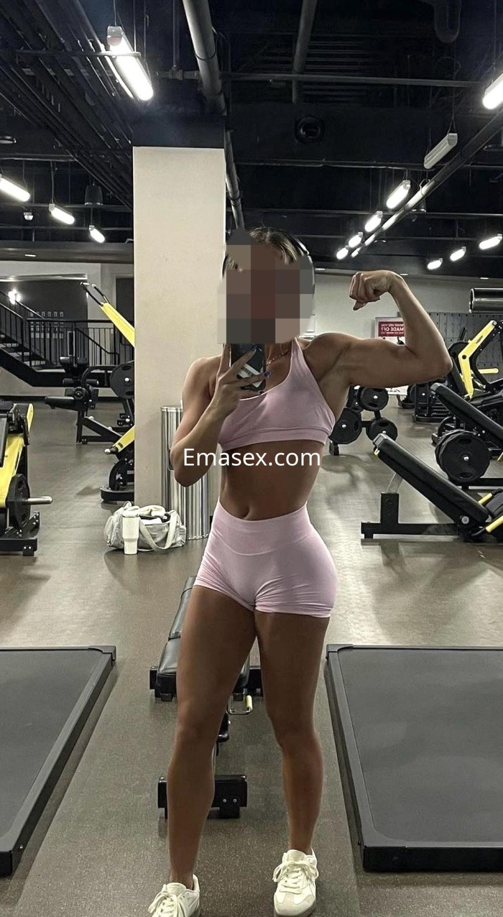Escort en Bilbao posando en un gimnasio con un conjunto deportivo rosa, flexionando el brazo y sonriendo.