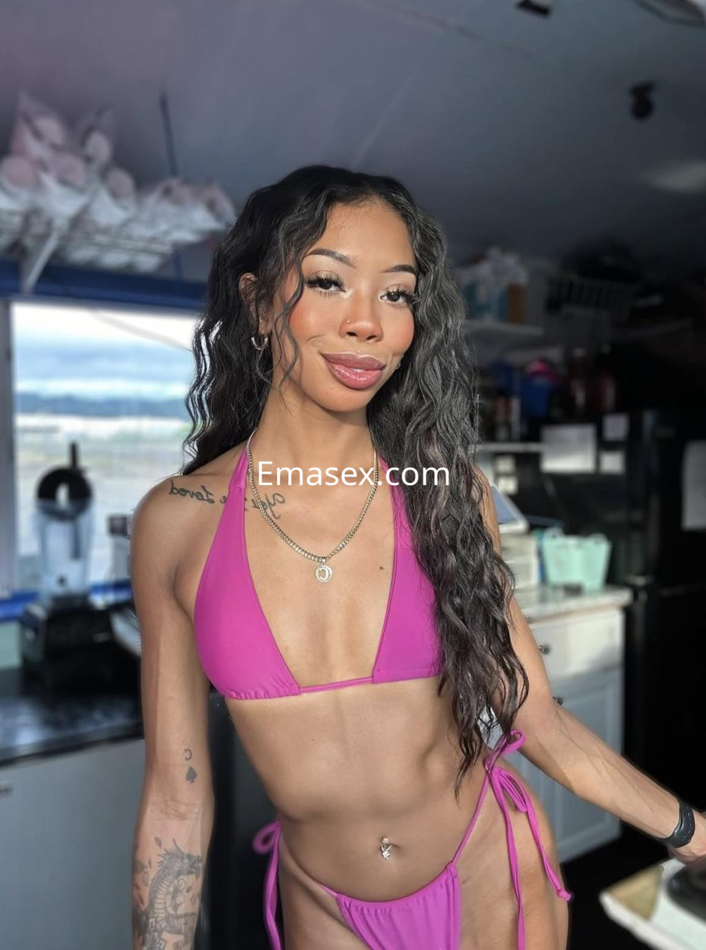 La escort en Irún lleva un bikini rosa, sonríe mientras posa en un ambiente iluminado con una cocina al fondo.