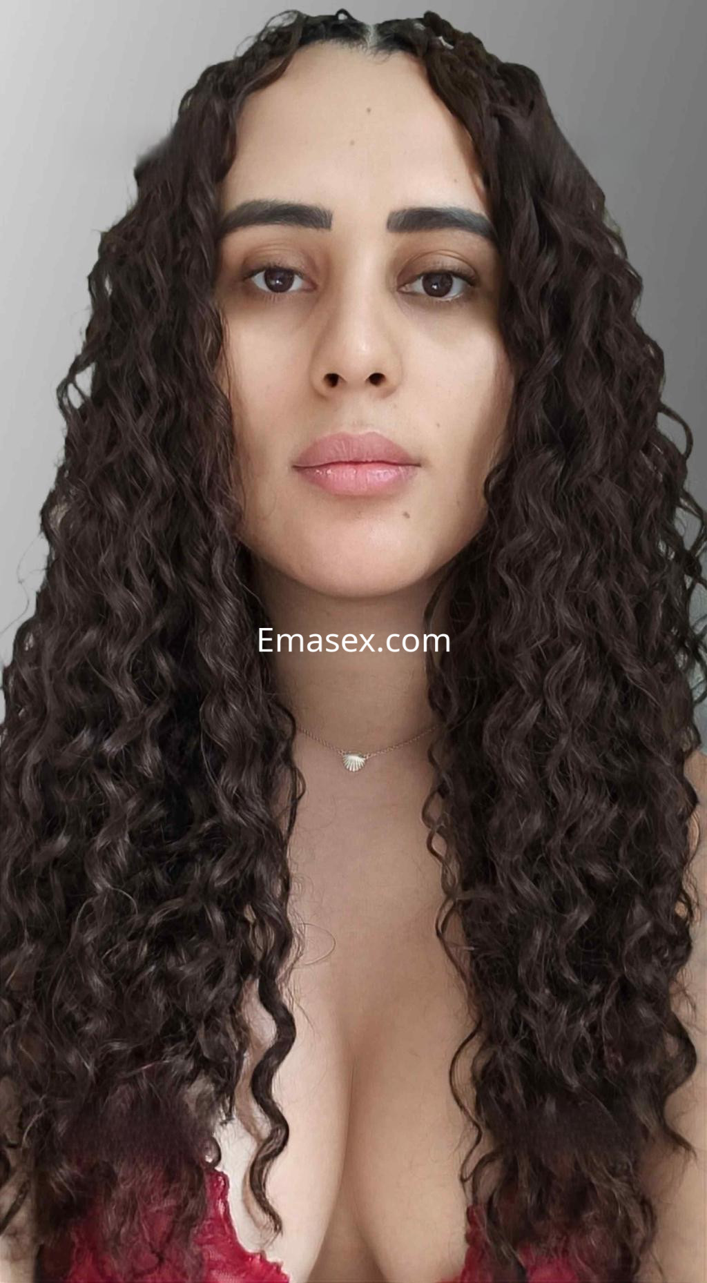Puta en Madrid con cabello rizado, posando de frente con un escote rojo y expresión cautivadora.
