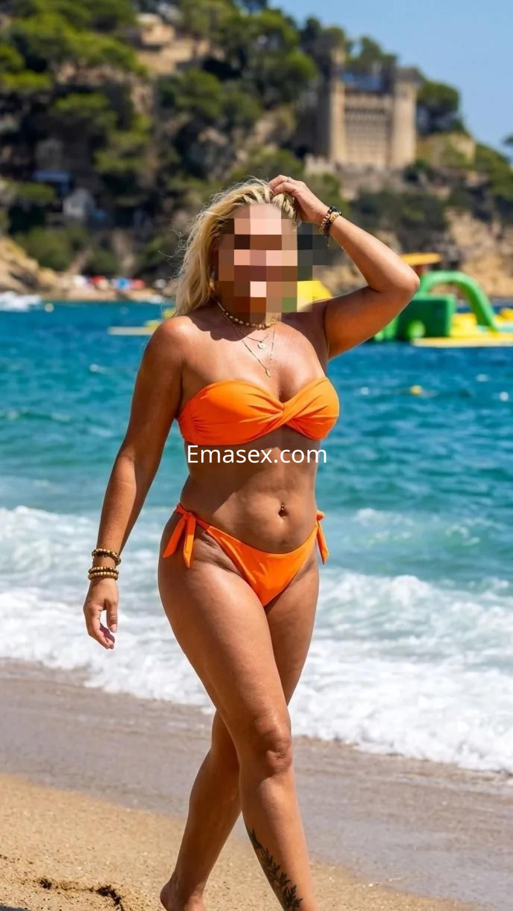 Escort en Girona caminando por la playa con un bikini naranja, luciendo un cabello rubio y un tatuaje en la pierna.