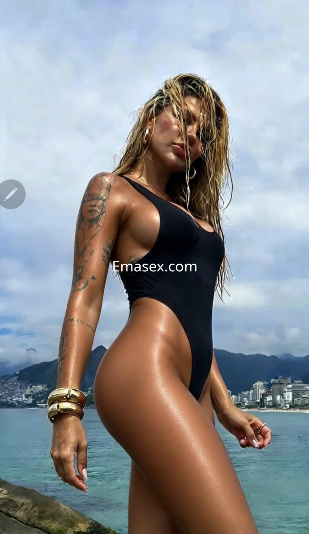 Puta en Logroño, con cabello rubio y húmedo, vistiendo un bañador negro y posando en la playa con tatuajes visibles.