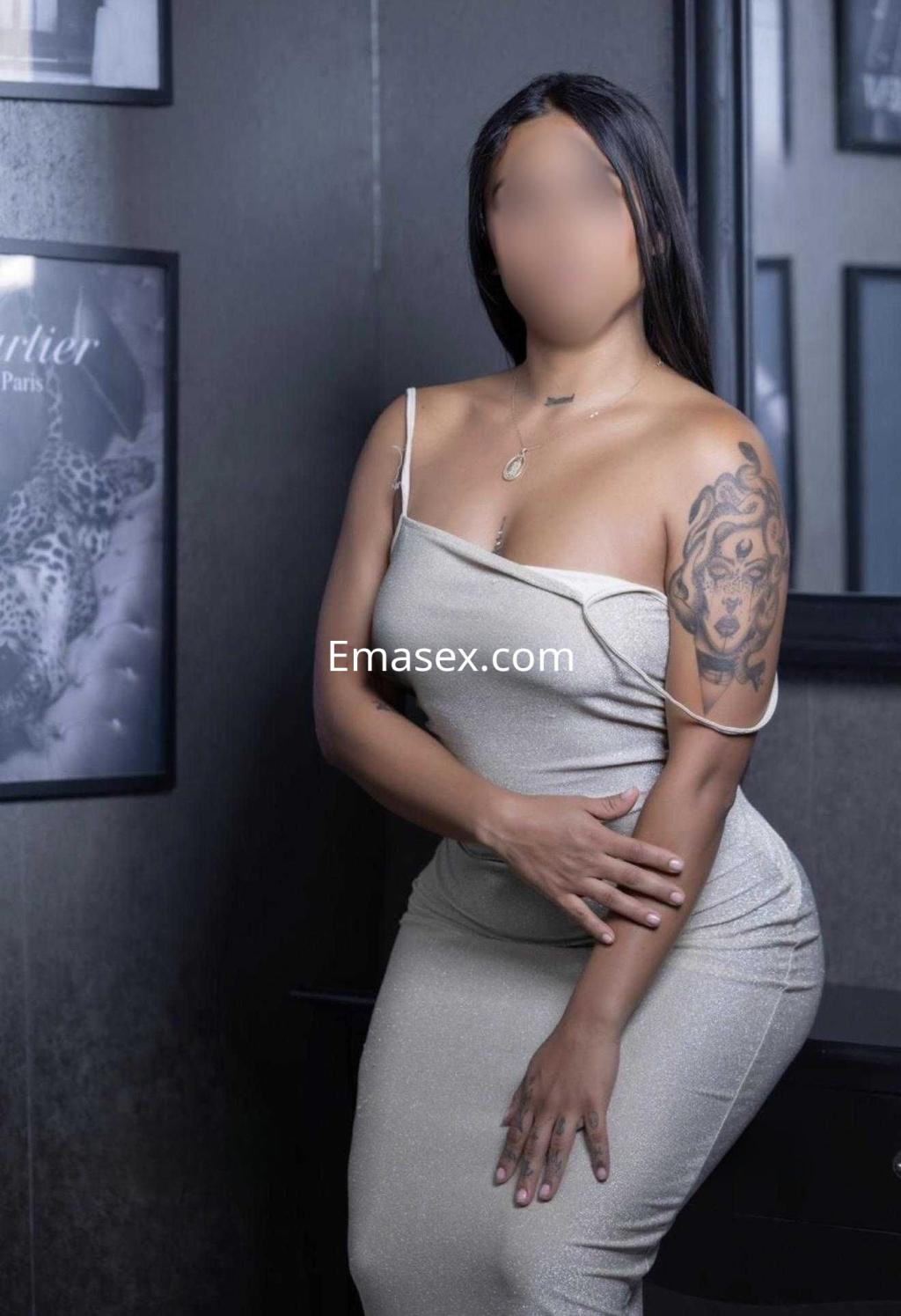 Puta posando en Barcelona con vestido ajustado plateado, cabello largo y lacio, y un tatuaje visible en el brazo.