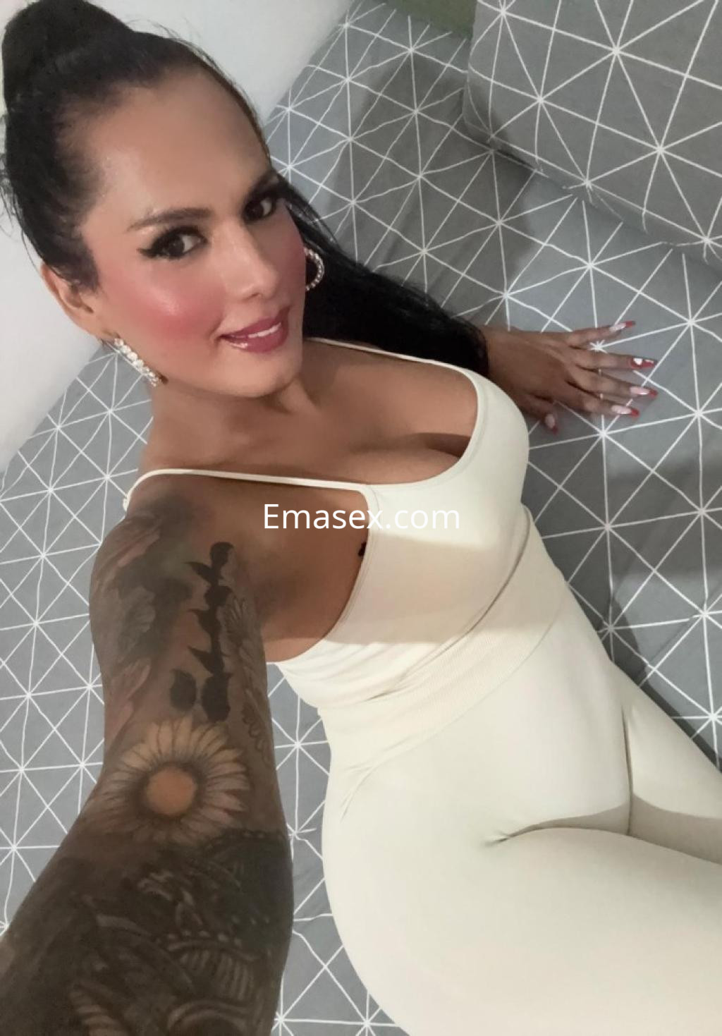 Escort en Tarragona posa con un mono blanco en un sofá gris, mostrando un tatuaje en el brazo y una sonrisa.