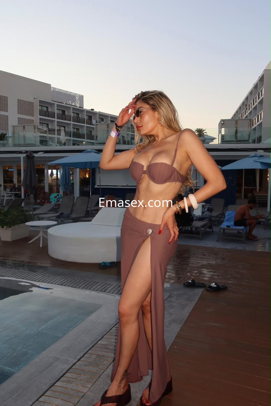Escort en Vigo luciendo un bikini marrón con una falda larga, posando junto a una piscina al atardecer.
