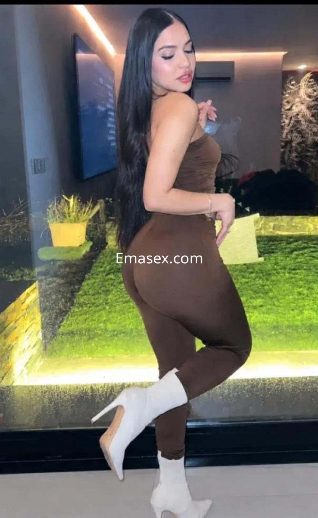 Escort en Barcelona posando de perfil, vistiendo un conjunto ceñido marrón y tacones blancos en un ambiente moderno.