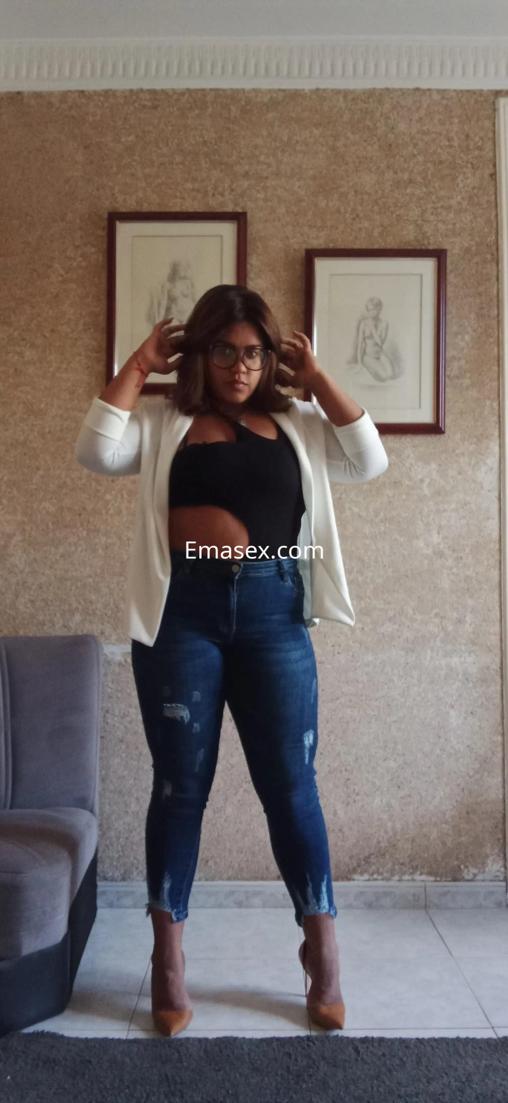 Escort en Barcelona, viste una blusa negra ajustada y chaqueta blanca, con jeans ajustados y tacones altos.