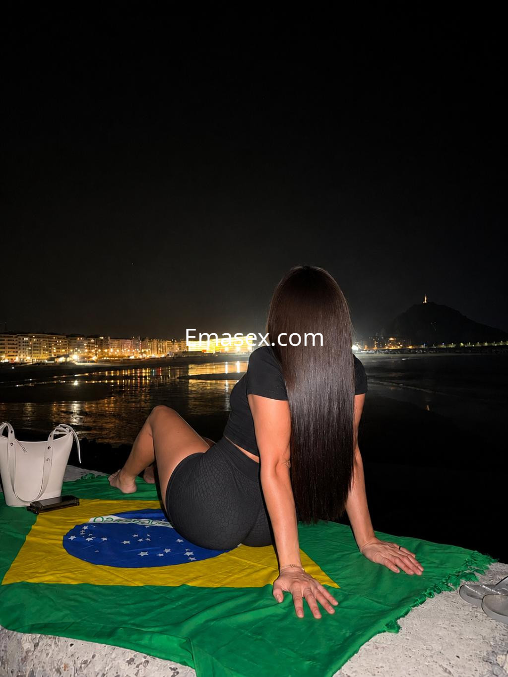La puta está sentada sobre una bandera de Brasil, luciendo un top negro y una falda corta, con la noche de Donosti de fondo.