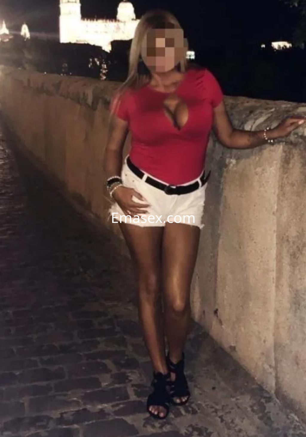 Escort en Gijón, con blusa roja ajustada y pantalones cortos blancos, posando en una calle iluminada.