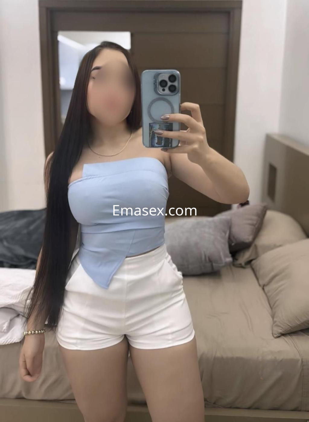 Escort en Huelva con blusa azul sin mangas y pantalones cortos blancos, posando frente a un espejo.