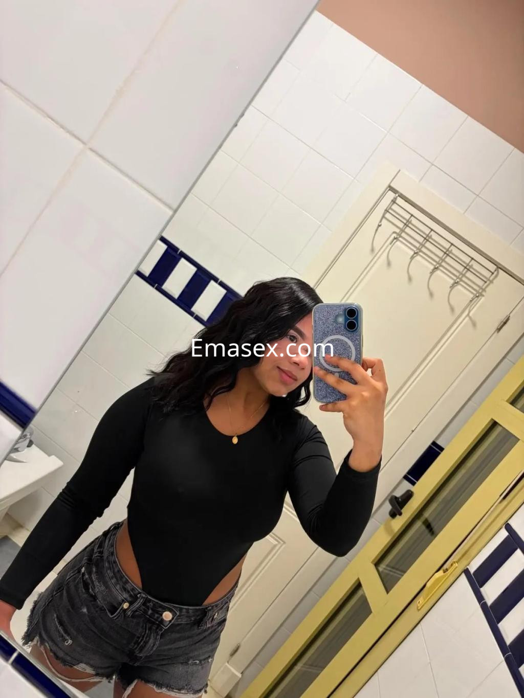 Escort en Córdoba con body negro y pantalones cortos de mezclilla, tomando un autorretrato en un baño.