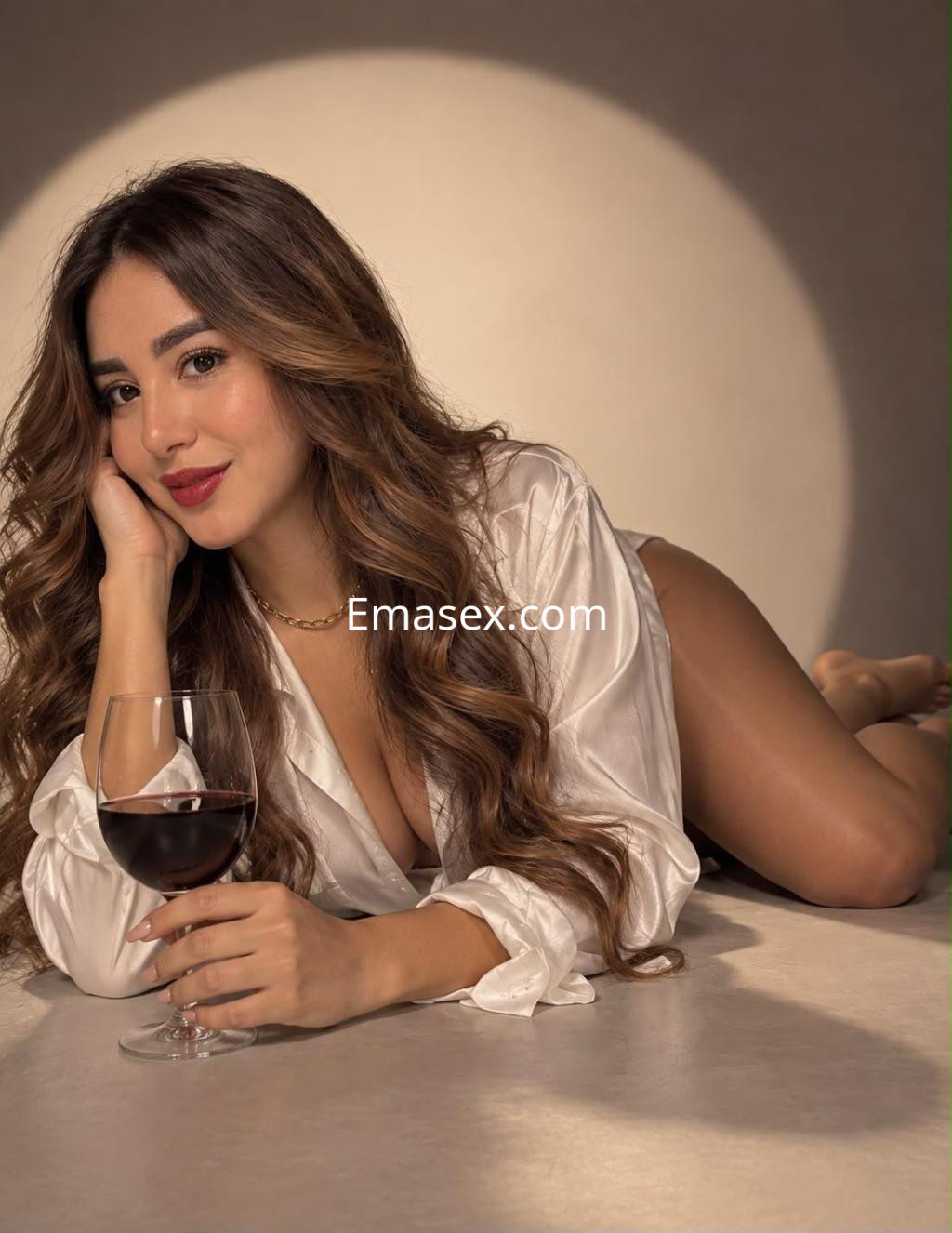 Puta en Madrid, recostada sobre una superficie clara, vistiendo una blusa blanca y sujetando una copa de vino.