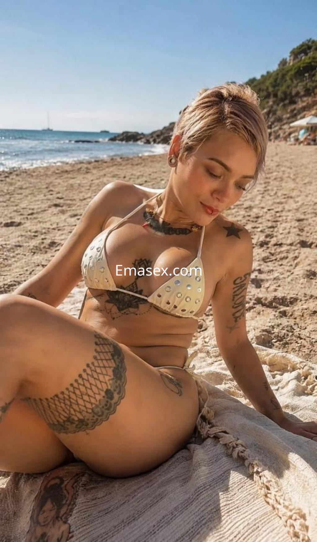 Escort en Huelva con cabello corto y rubio, posando en la playa con bikini blanco y tatuajes visibles.