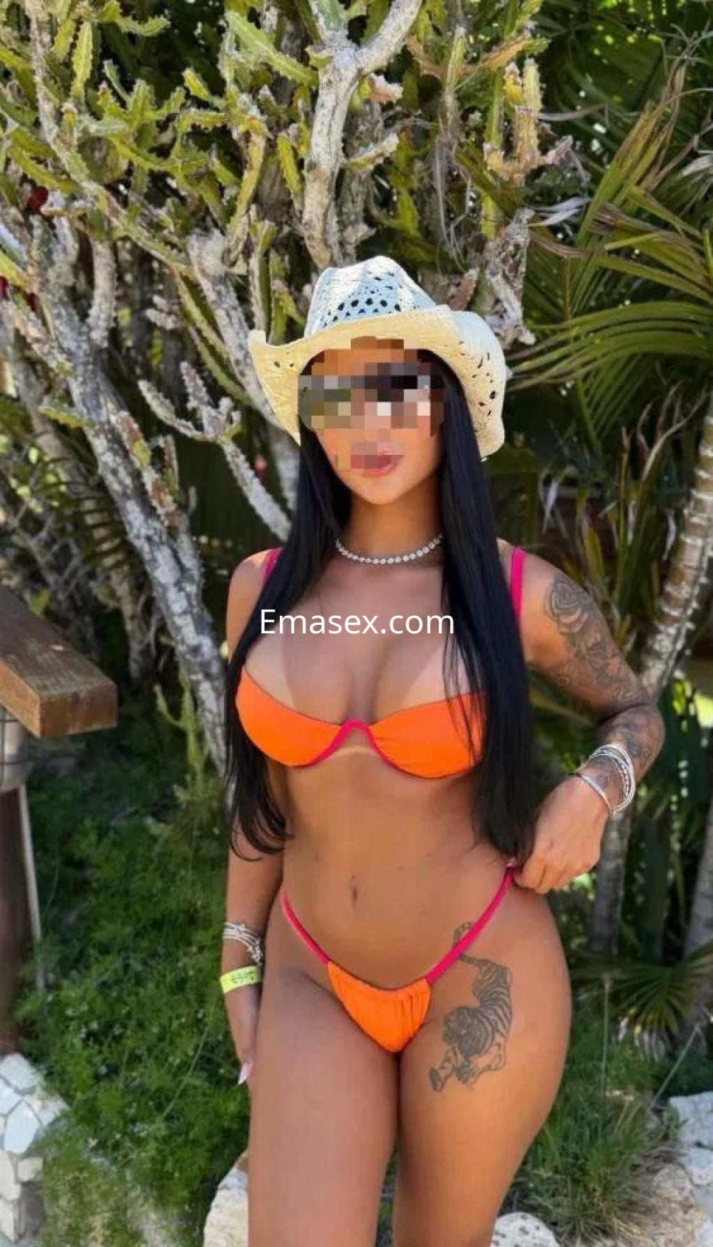 Escort en Pontevedra luciendo un bikini naranja con detalles en rosa y un sombrero blanco, posando en un entorno tropical.