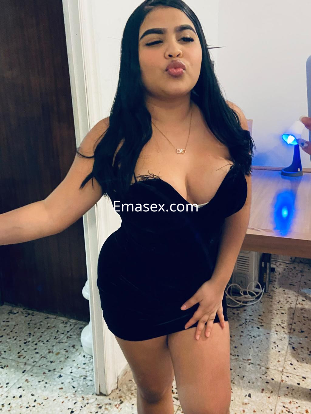 Escort en Almería posando con un vestido negro ajustado, con cabello largo y lacio, en un entorno interior.