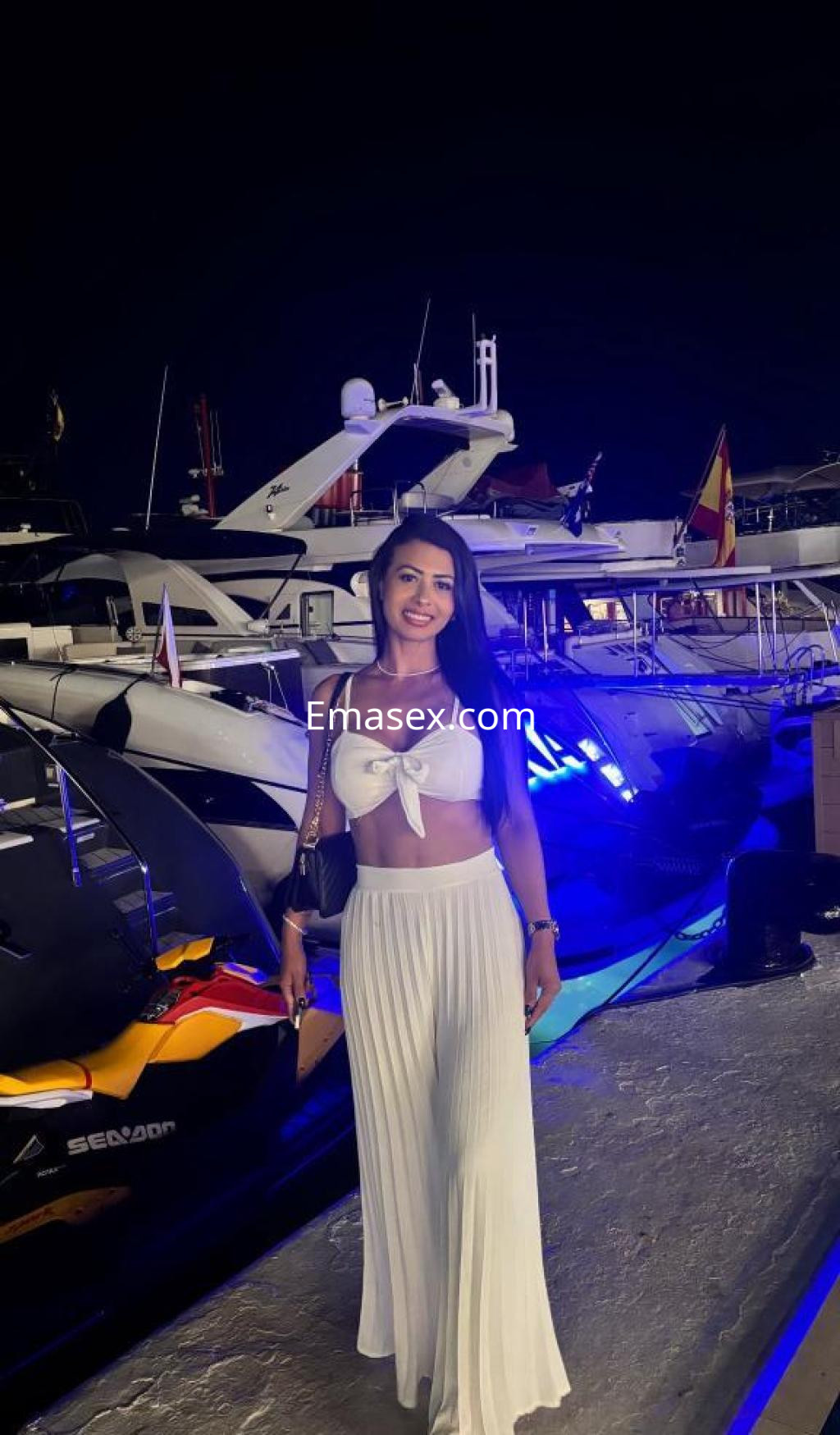 Escort en Lugo, posando con un top blanco y falda larga, rodeada de yates bajo luces nocturnas.