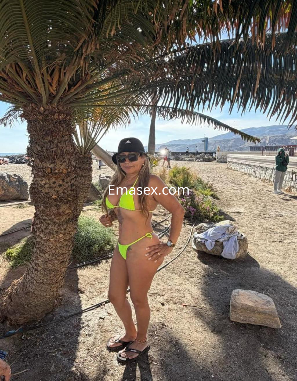 Puta con bikini amarillo neón y gorra negra, posando en la playa de Almería, rodeada de palmeras.