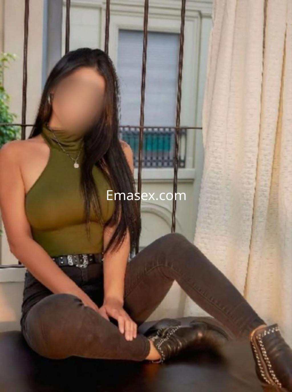 Puta en Valladolid con cabello lacio, usando un top de cuello alto verde y jeans oscuros, sentada en un sillón.