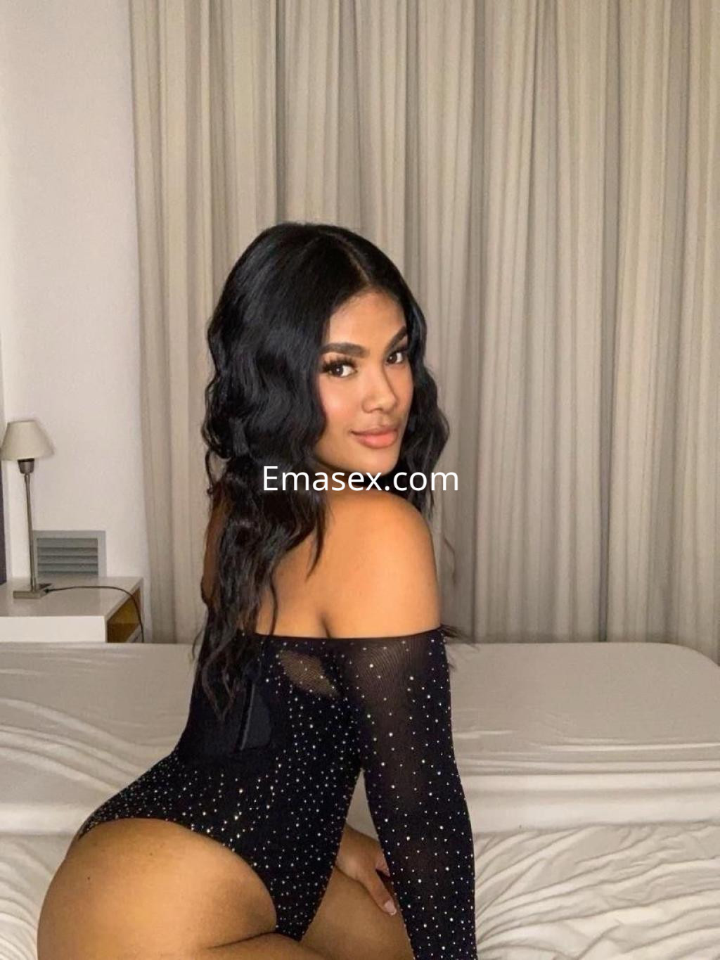 Escort en Murcia con un body negro con brillo, posando de perfil en una habitación con cortinas claras.
