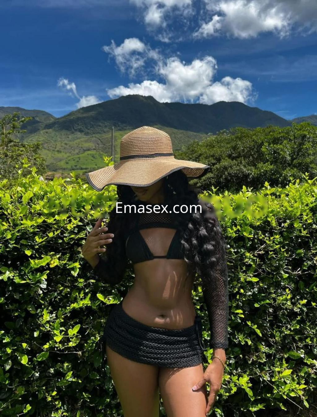 Escort en Girona con cabello largo y rizado, vistiendo un bikini negro y un sombrero grande, posando en un entorno natural.
