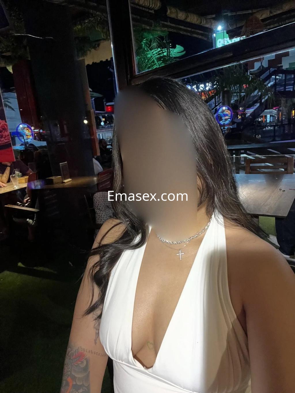 Puta en Murcia luciendo un vestido blanco con escote, acompañada de joyería brillante en un ambiente nocturno.
