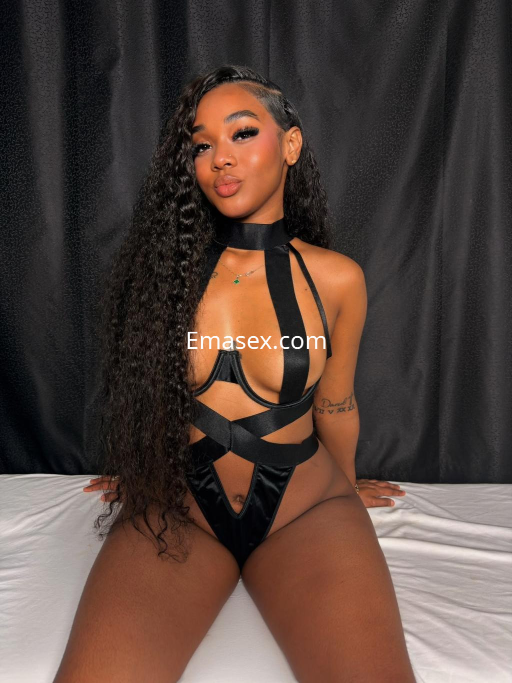 Escort en Madrid con cabello largo y rizado, vistiendo un conjunto negro de lencería con tiras, posando sentada.
