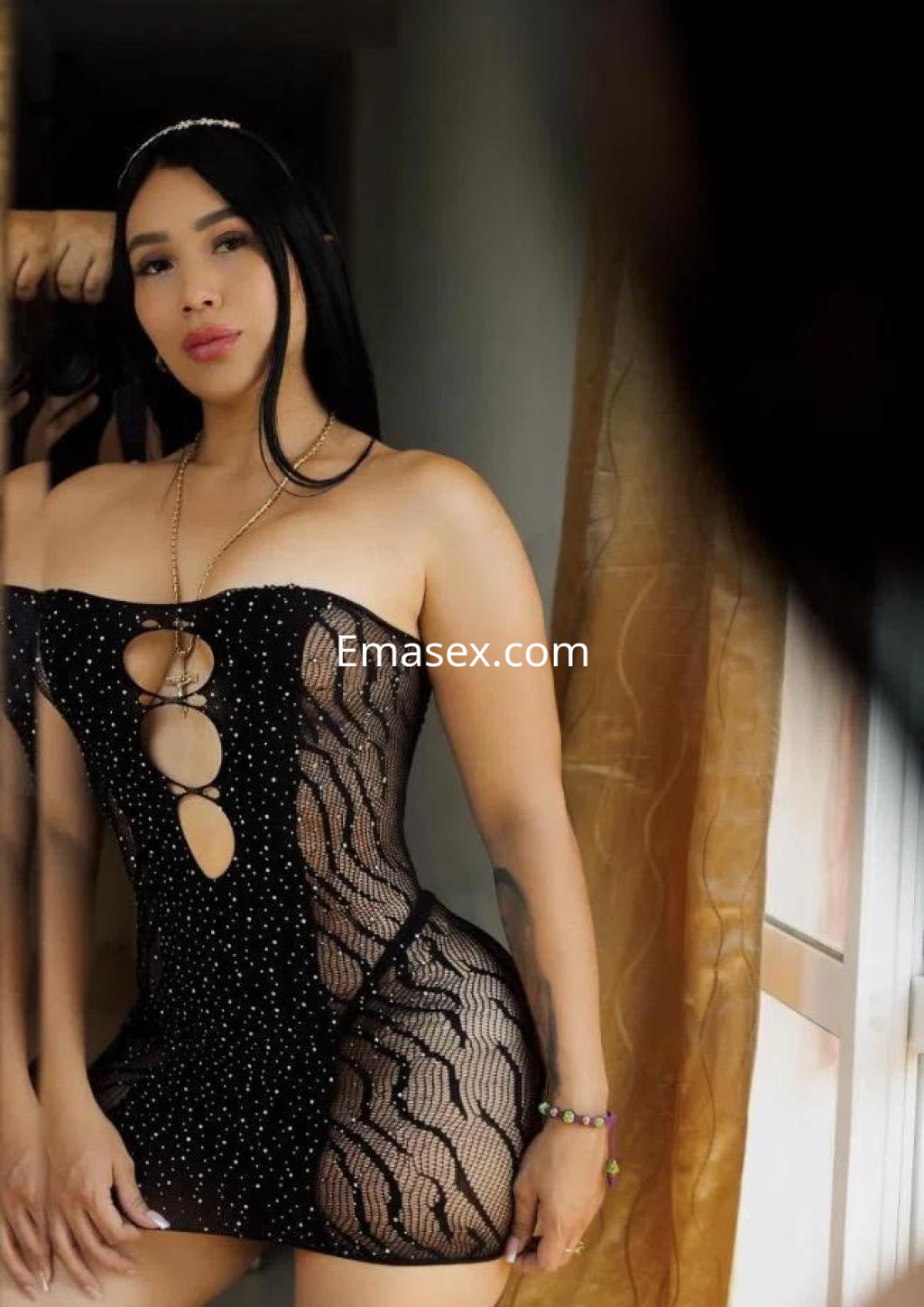 Escort en Sevilla con vestido negro ajustado, detalles brillantes y cabello largo, posando frente a un espejo.