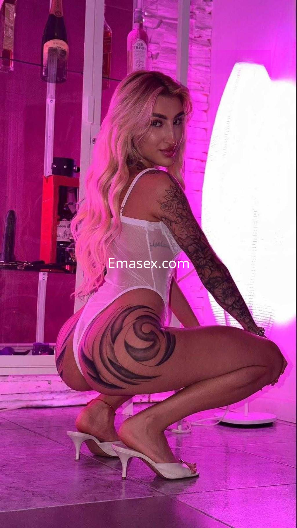 Escort en Granada con cabello largo y rubio, vestida con un body blanco y tacones, posando en un ambiente iluminado.